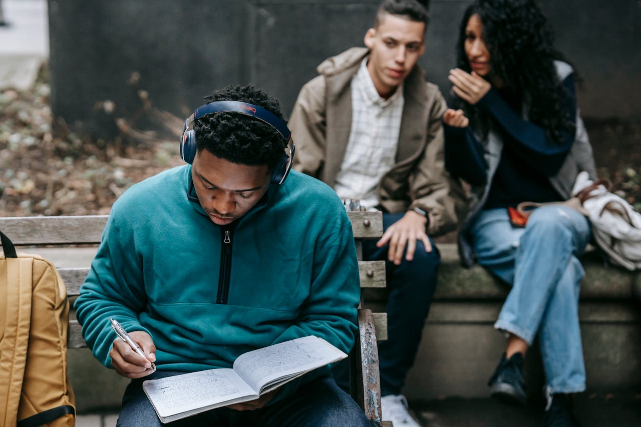 Crop young ethnic man and woman sitting on bench and gossiping about concentrated African American male student listening to music in headphone and doing homework assignment