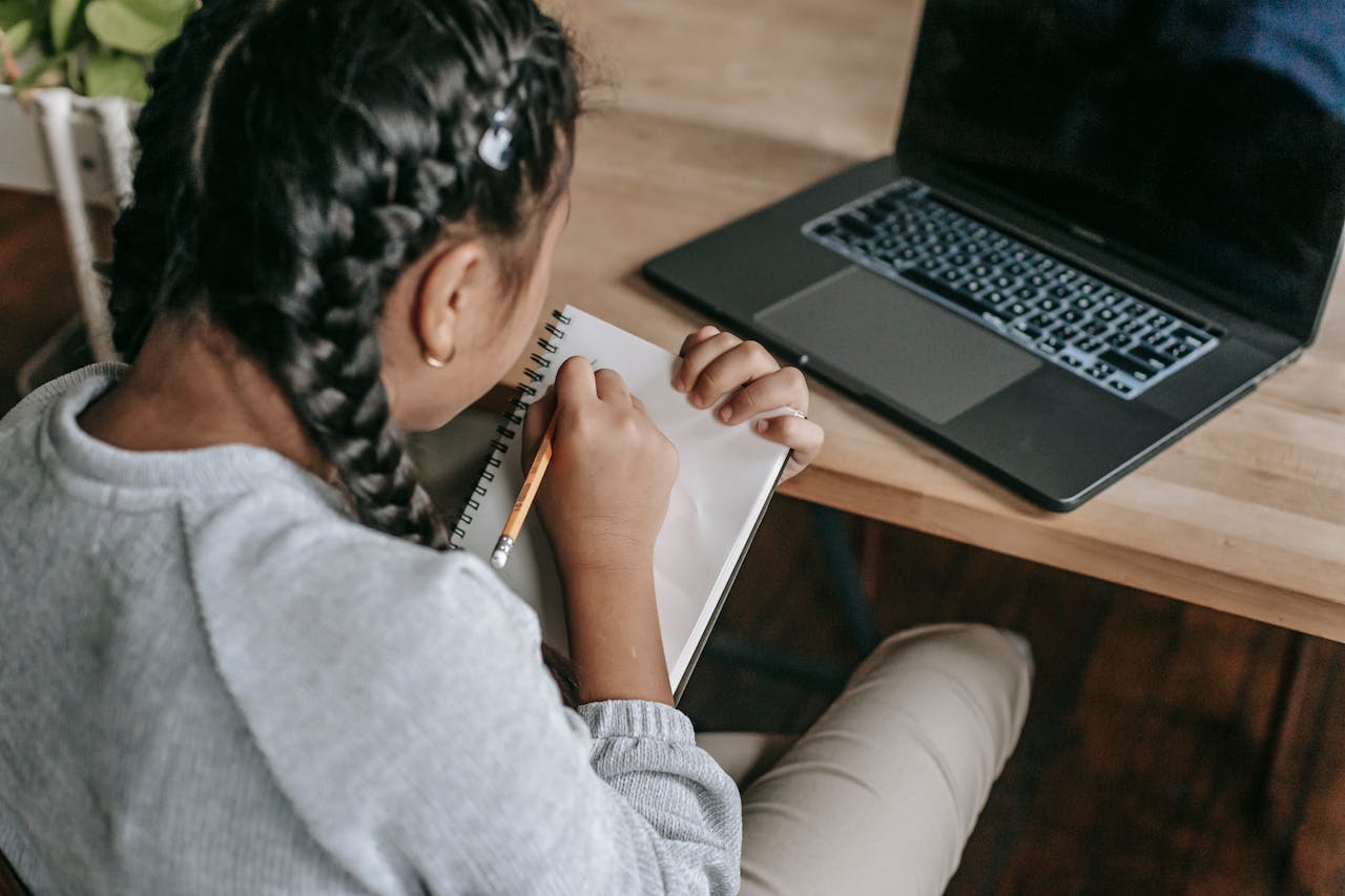 Young girl writing in a notebook with a laptop, showcasing focused learning.